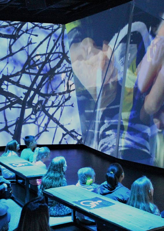 A group of children sit in a small theatre to watch wall-to-wall screens with Canadian scenery.