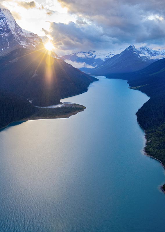 An aerial view of a large lake surrounded by forested mountains, with the Sun shining.